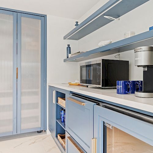 A modern kitchen with blue cabinets, a microwave, a coffee maker, mugs, and shelves containing various items against a white background.