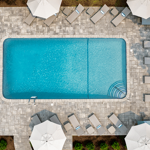 The image shows a rectangular swimming pool surrounded by lounge chairs, umbrellas, plants, and a paved area in a landscaped outdoor setting.