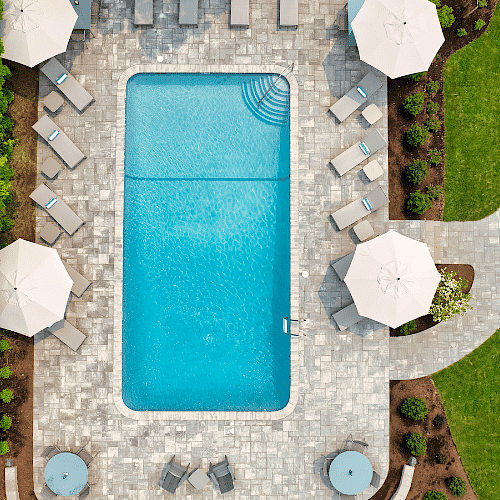 An aerial view of a rectangular swimming pool with surrounding lounge chairs and umbrellas, nestled in a landscaped area with greenery and pathways.