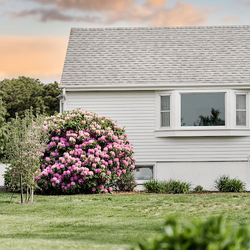 A white house with a shingled roof, a large bush with pink flowers, green lawn, and flowering plants in the foreground, under a partly cloudy sky.