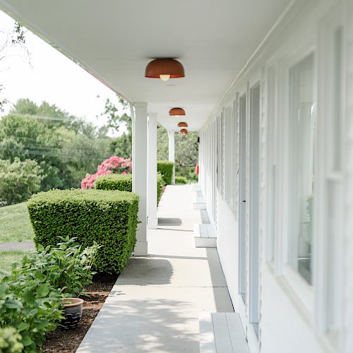 The image shows a long, covered walkway with white walls and windows on one side, and a neatly trimmed hedge and garden on the other side.