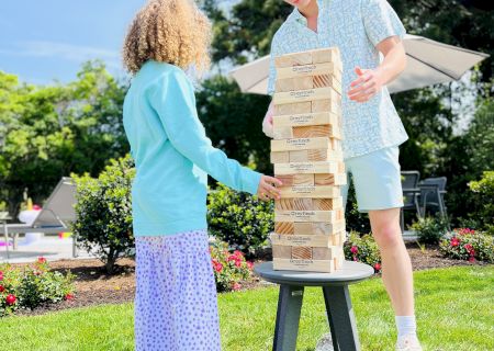 Two people are playing a game of giant Jenga on a stool outdoors in a garden, with a sunny sky and trees in the background.