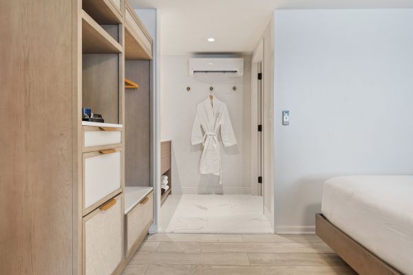 A modern, minimalist hotel room featuring a neatly organized closet, a bathrobe on a hook, and a neatly made bed in a light wood and white theme.