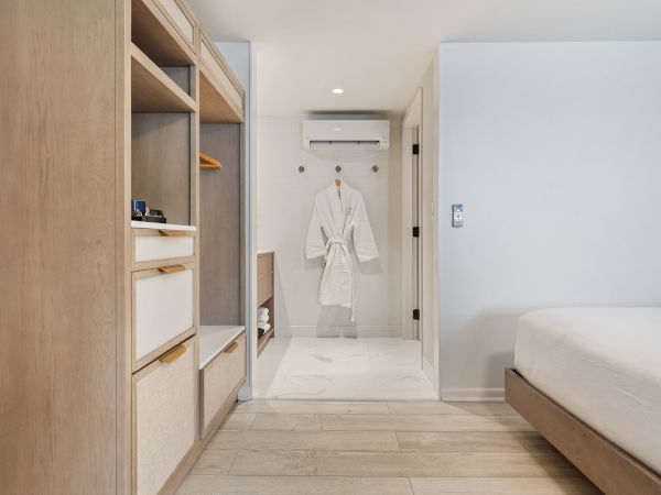 A modern, minimalist hotel room featuring a neatly organized closet, a bathrobe on a hook, and a neatly made bed in a light wood and white theme.