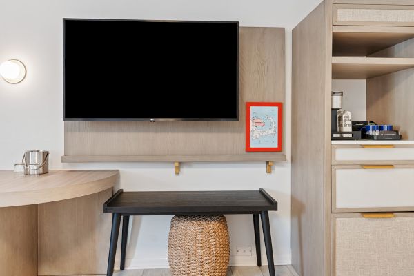 The image shows a modern hotel room setup with a wall-mounted TV, a black desk with a wicker basket underneath, and a cabinet with a minibar.