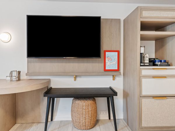 The image shows a modern hotel room setup with a wall-mounted TV, a black desk with a wicker basket underneath, and a cabinet with a minibar.