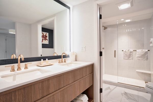 This image shows a modern bathroom with a double sink vanity, large mirror, wooden cabinets, and a glass-enclosed shower area with white towels.