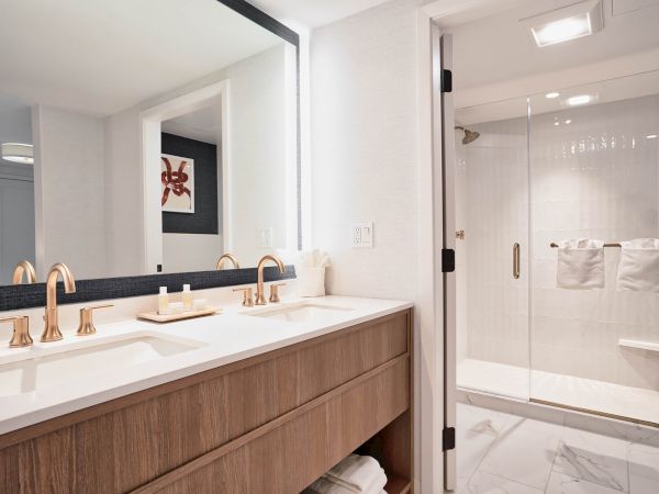 This image shows a modern bathroom with a double sink vanity, large mirror, wooden cabinets, and a glass-enclosed shower area with white towels.