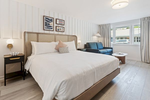 A neat bedroom with a bed, nightstands, a sofa, a coffee table, lamps, and framed art on the wall. A window shows an exterior view.