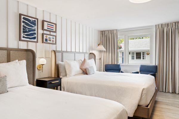 The image shows a clean, modern hotel room with two double beds, artwork on the walls, a window with curtains, and a sitting area with two blue chairs.