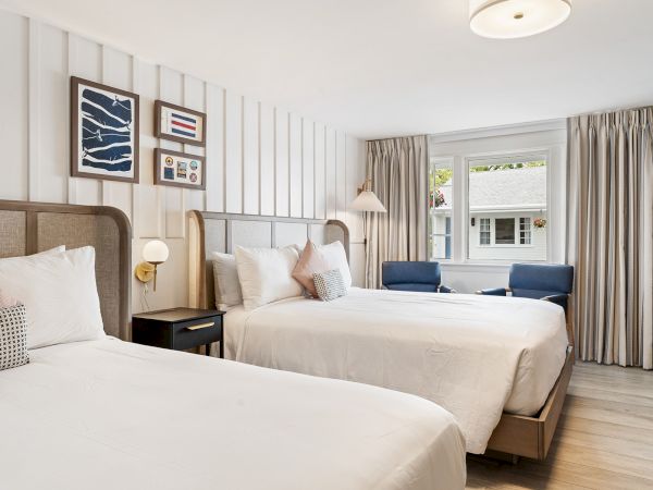 The image shows a clean, modern hotel room with two double beds, artwork on the walls, a window with curtains, and a sitting area with two blue chairs.