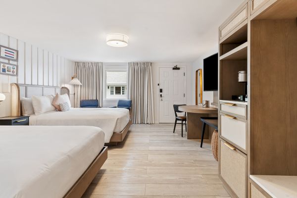 A modern hotel room with two beds, a desk, a chair, a TV on the wall, a storage unit, and a large window with curtains in the background.