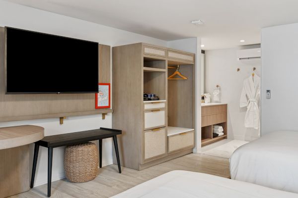 This image shows a modern hotel room with two beds, a TV, a desk, an open wardrobe, robes, a bathroom area, and minimalistic decor.