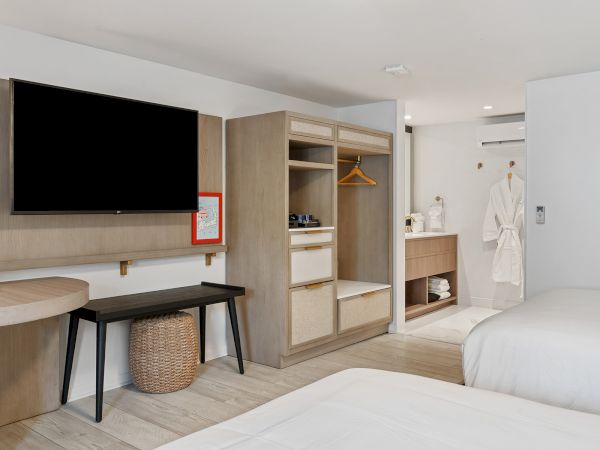 This image shows a modern hotel room with two beds, a TV, a desk, an open wardrobe, robes, a bathroom area, and minimalistic decor.