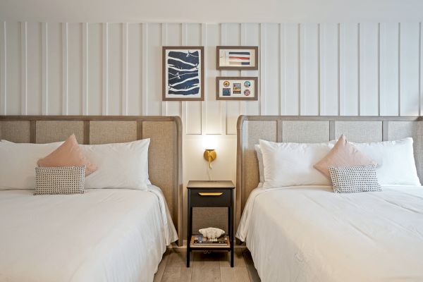 The image depicts a neatly arranged bedroom with two double beds, a nightstand with a lamp and books, and framed artwork above the headboards.