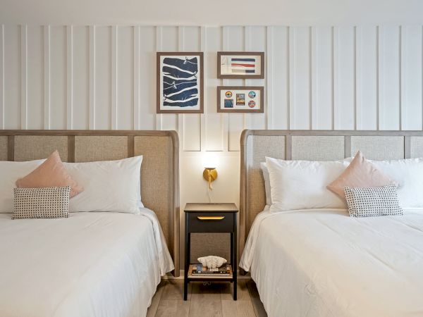 The image depicts a neatly arranged bedroom with two double beds, a nightstand with a lamp and books, and framed artwork above the headboards.
