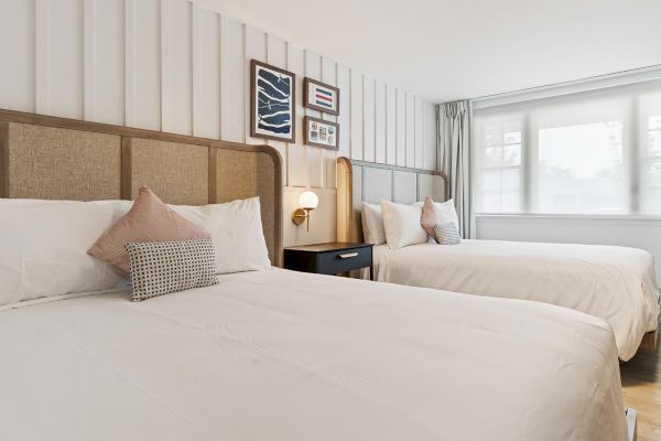 The image shows a well-lit bedroom with two neatly made beds, a nightstand with a lamp between them, and framed artwork on the wall.