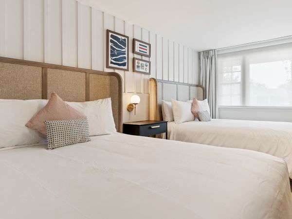 The image shows a well-lit bedroom with two neatly made beds, a nightstand with a lamp between them, and framed artwork on the wall.