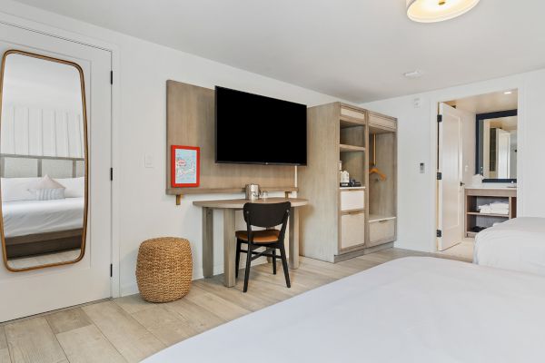 The image shows a modern hotel room with a bed, a TV, a desk, a chair, a woven basket, and a bathroom.