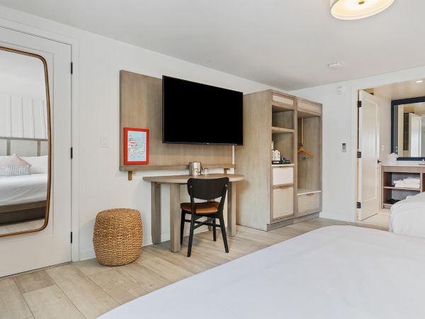 The image shows a modern hotel room with a bed, a TV, a desk, a chair, a woven basket, and a bathroom.
