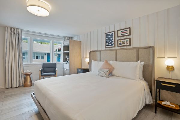 This is a bright, modern bedroom featuring a large bed, wall art, nightstand with light, a chair by the window, and wooden flooring.