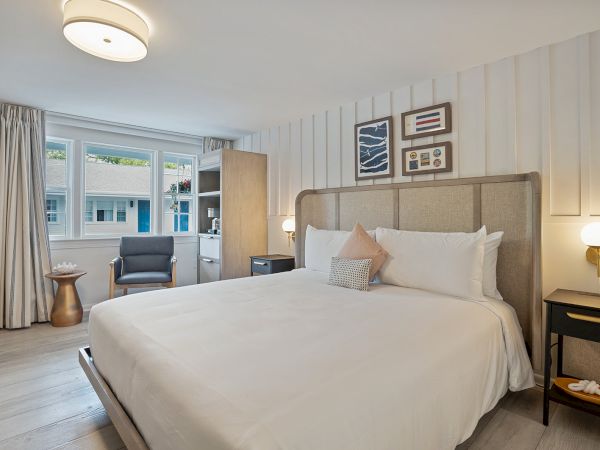 This is a bright, modern bedroom featuring a large bed, wall art, nightstand with light, a chair by the window, and wooden flooring.