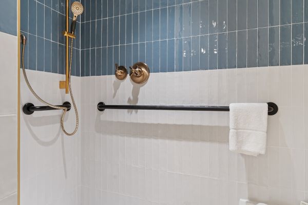 The image shows a modern shower with blue and white tiles, a handheld showerhead, grab bars, and a towel hanging on a bar.