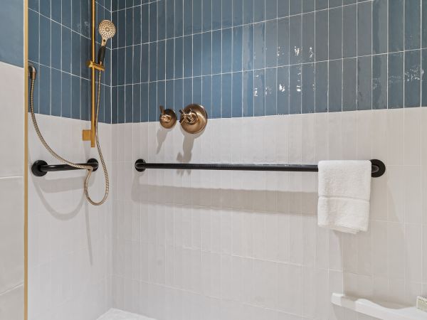 The image shows a modern shower with blue and white tiles, a handheld showerhead, grab bars, and a towel hanging on a bar.