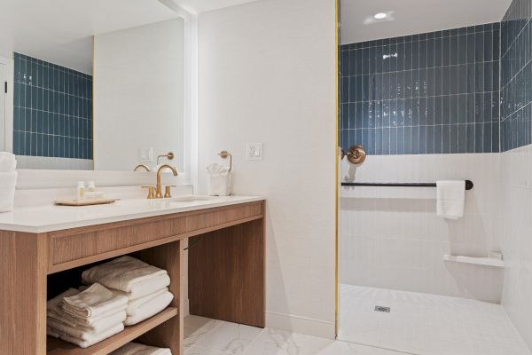 This image features a modern bathroom with a walk-in shower, a large mirror, a wooden vanity with a countertop, and neatly stacked towels.