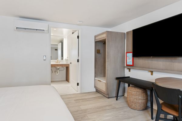 The image shows a modern hotel room with a bed, flat-screen TV, desk, chair, basket, and an open door leading to the bathroom.