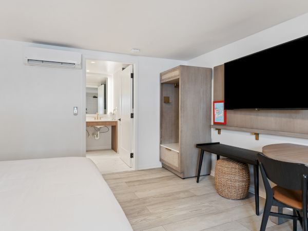 The image shows a modern hotel room with a bed, flat-screen TV, desk, chair, basket, and an open door leading to the bathroom.