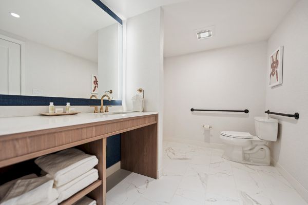 The image shows a modern bathroom with a large mirror, marble floor, a white toilet, towel storage beneath a sink counter, and minimalist wall art.