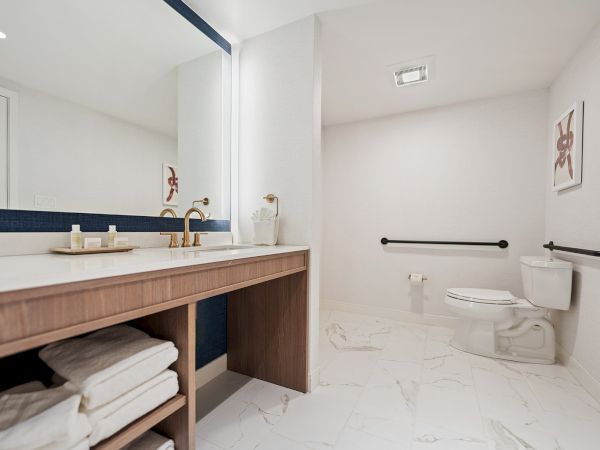 The image shows a modern bathroom with a large mirror, marble floor, a white toilet, towel storage beneath a sink counter, and minimalist wall art.