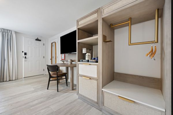 A modern hotel room with a TV, desk, chair, and partial view of the wardrobe area with hangers. Light wooden flooring and a white door.