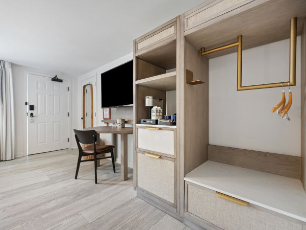 A modern hotel room with a TV, desk, chair, and partial view of the wardrobe area with hangers. Light wooden flooring and a white door.