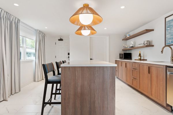 A modern kitchen with wooden cabinets, an island with bar stools, pendant lights, and stainless steel appliances. It has white walls and large windows.