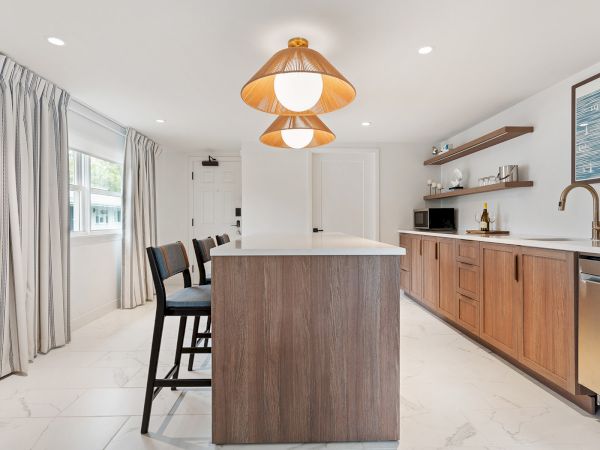 A modern kitchen with wooden cabinets, an island with bar stools, pendant lights, and stainless steel appliances. It has white walls and large windows.