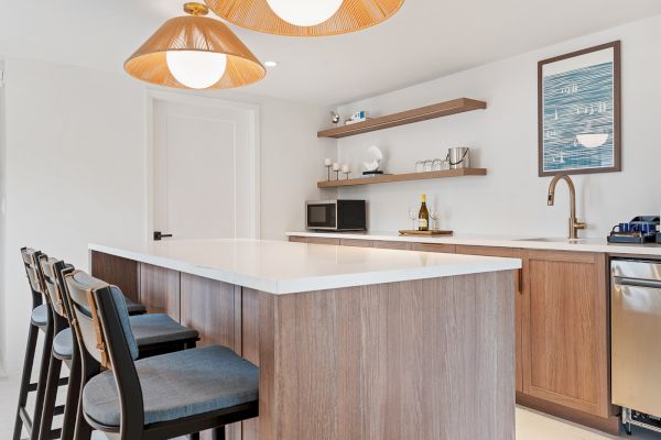 A modern kitchen with a large island, wooden cabinets, pendant lights, open shelves, and three bar stools.