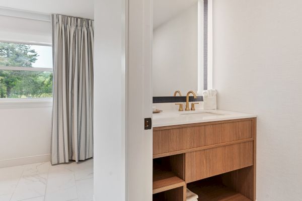 The image shows a bathroom with a wooden vanity, gold faucet, mirror, and a stack of towels. To the left, a window with curtains is partially visible.