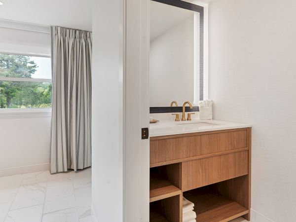 The image shows a bathroom with a wooden vanity, gold faucet, mirror, and a stack of towels. To the left, a window with curtains is partially visible.
