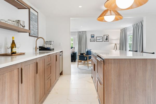 A modern kitchen with wooden cabinets, a wine bottle, glasses, and shelves. Pendant lights hang above the island. The kitchen opens to a living area.