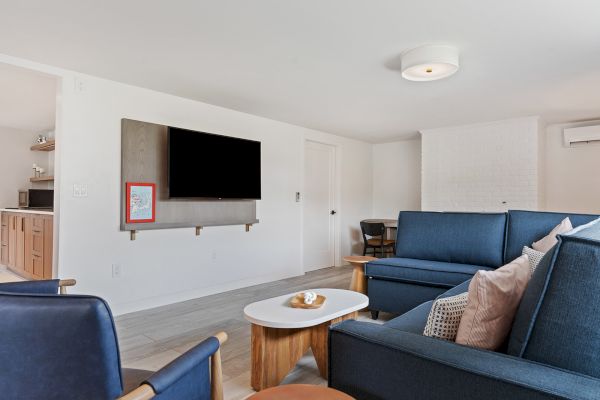 The image shows a living room with blue couches, a wall-mounted TV, and a small coffee table. There is a kitchen visible in the background.