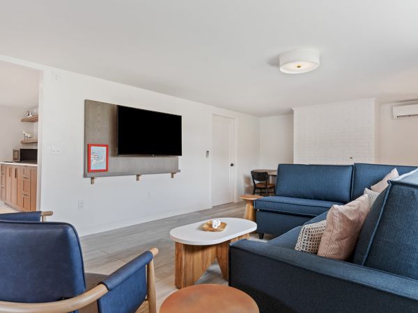 The image shows a living room with blue couches, a wall-mounted TV, and a small coffee table. There is a kitchen visible in the background.