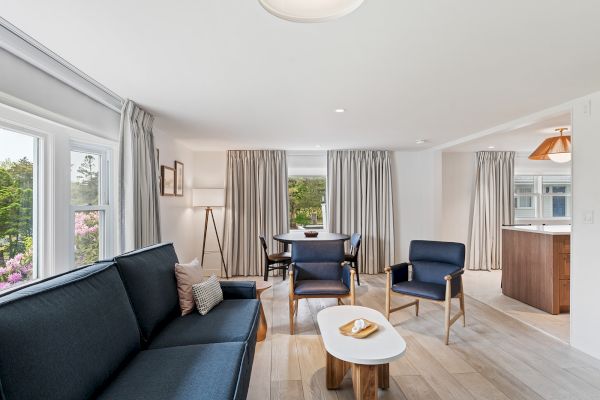 A bright, modern living room with a blue couch, two blue armchairs, a white coffee table, and large windows with gray curtains.