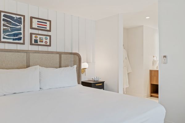 A modern hotel room with a neatly made bed, wall art, bedside table with lamp, and a view into a bathroom with robes hanging.