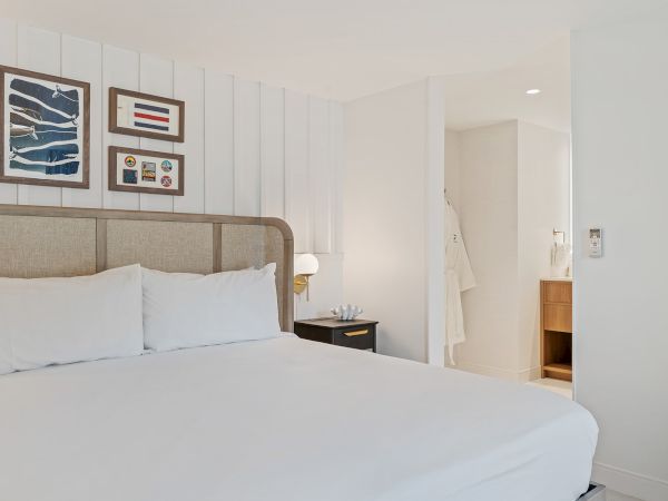 A modern hotel room with a neatly made bed, wall art, bedside table with lamp, and a view into a bathroom with robes hanging.