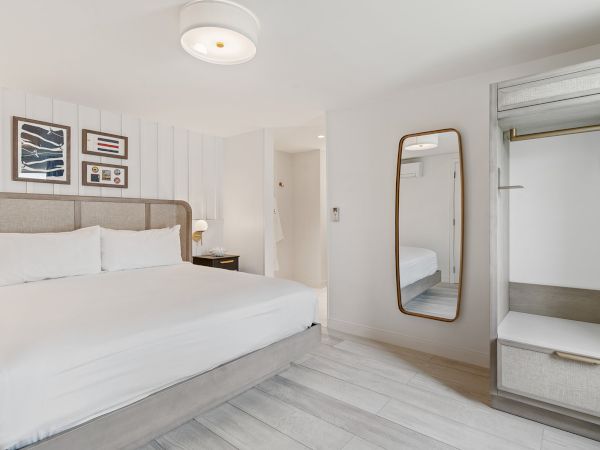 A modern, minimalistic bedroom with a large bed, wall art, a bedside table, and a full-length mirror near an open wardrobe.