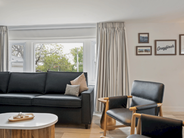 A modern living room features a black sofa, armchairs, a coffee table, wall art, and large windows with light curtains, creating a cozy ambiance.