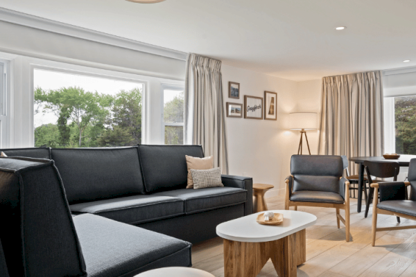 This image shows a modern living room with a black couch, wooden furniture, a dining area, large windows, framed pictures on the wall, and a lamp.