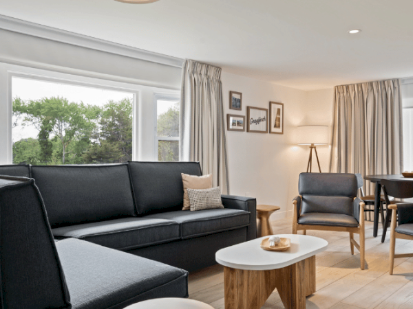 This image shows a modern living room with a black couch, wooden furniture, a dining area, large windows, framed pictures on the wall, and a lamp.